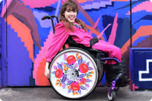 On a outdoor colouful backdrop a woman in a wheelchair shows a floral designed wheelchair cover with red and yellow flowers and purple leaves